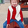 child, playful, outdoor, picnic_table, wooden, casual_clothing, vest, jeans, sneakers, sunny, tongue_out, squinting, funny_face, smiling, daylight, concrete, building_wall, window, shadow, person