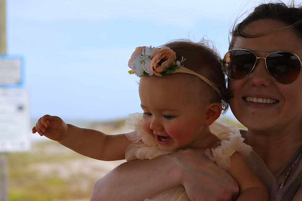 Amelia is registered to the contest to win money with this photo: baby, child, woman, sunglasses, flower_headband, smile, outdoor, sunlight, happy, portrait, holding, arm, dress, skin, face, person, head, nature, daylight, background_blur