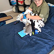 Speedy is registered to the contest to win money with this photo: child, bed, blanket, kitten, tablet, glasses, plush_toys, shelf, blue_bedspread, remote_control, floor, room, curly_hair, smiling, indoor, clothing, toy, playful, pet, happy