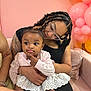 child, woman, baby, braids, glasses, pink_sweater, white_skirt, balloons, pink_wall, sofa, couch, tattoo, bracelet, happy, indoor, celebration, family, cute, portrait, affection