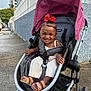 Elyana is registered to the contest to win money with this photo: toddler, child, stroller, smile, red_bow, sandals, urban, sidewalk, outdoor, happy, baby, clothing, footwear, fence, building, concrete, sunlight, person, portrait, casual