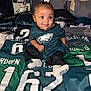 baby, child, football_jersey, philadelphia_eagles, sports_fan, bed, pillow, blanket, clothing, cute, indoors, portrait, smiling, socks, hair_bow, team_spirit, memorabilia, fan_gear, person, home