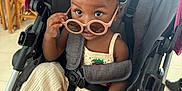 Elyana is registered to the contest to win money with this photo: toddler, child, stroller, sunglasses, red_bow, clothing, footwear, indoor, furniture, curious, baby, person, seatbelt, cute, expression, sandals, face, headwear, accessories, floor