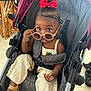 toddler, child, stroller, sunglasses, red_bow, clothing, footwear, indoor, furniture, curious, baby, person, seatbelt, cute, expression, sandals, face, headwear, accessories, floor