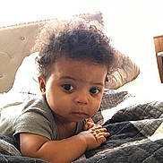 Jabari joined the competition — help win amazing prizes! baby, infant, child, curly_hair, bed, blanket, pillow, indoor, portrait, cute, looking, face, hands, cozy, young, person, clothing, sleeping_area, soft, expression