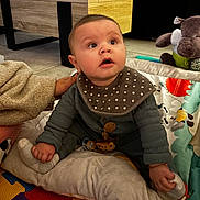 Eden a rejoint le concours — aidez-le/la à gagner de superbes lots ! baby, child, indoor, play_mat, bib, sock, hand, plush_toy, hippo, blanket, curious, floor, wooden_table, colorful, clothing, person, cute, expression, seated, toddler