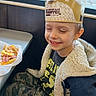 child, boy, smiling, restaurant, booth, crown, paper_crown, breakfast, bacon, eggs, food, dining, indoor, portrait, happy, seated, jacket, camouflage_pants, window, vehicle