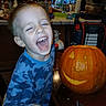 child, toddler, boy, laughing, smiling, face, portrait, pumpkin, jack_o_lantern, halloween, pumpkin_carving, carving_tool, kitchen, indoors, home, table, blue_shirt, wooden_cabinets, holiday, autumn