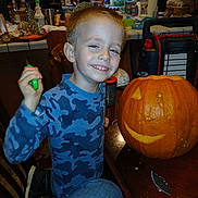 Ferris is registered to the contest to win money with this photo: child, boy, smiling, portrait, pumpkin, jack_o_lantern, halloween, carving, kitchen, indoors, table, camouflage_shirt, blue_clothing, hand_tool, crafting, food, chair, home, celebration, photography