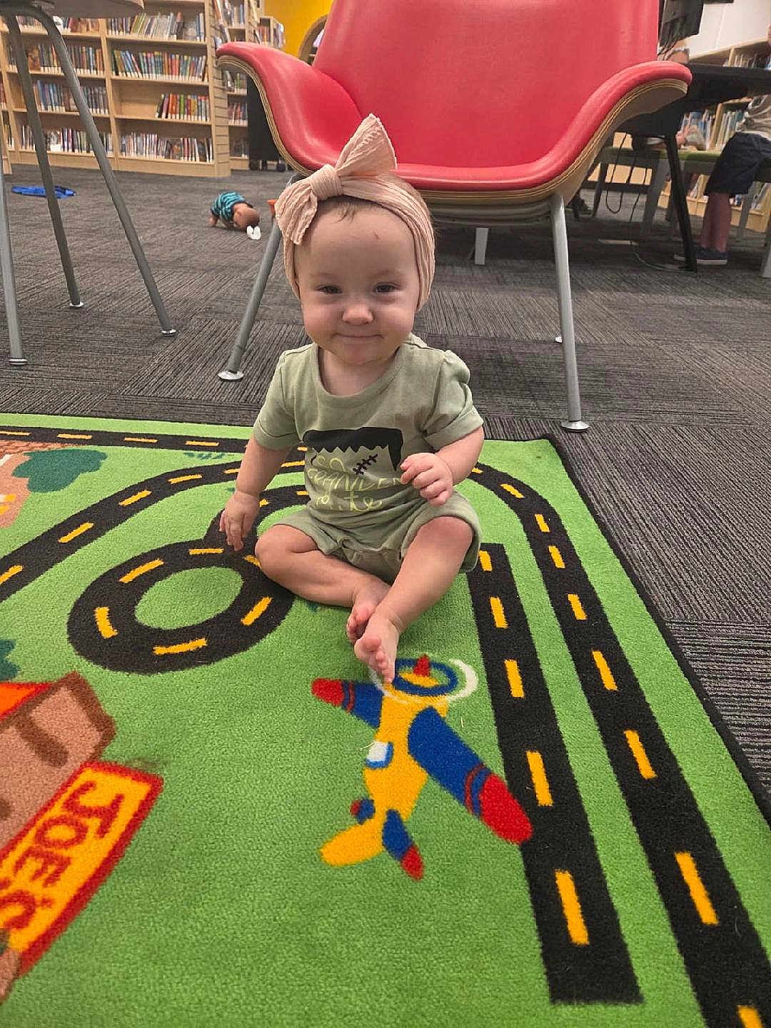 Charlotte Mae is registered to the contest to win money with this photo: baby, child, smiling, headband, bow, play_mat, colorful, airplane, road_pattern, library, bookshelf, carpet, floor, chair, indoor, person, sitting, cute, toddler, clothing