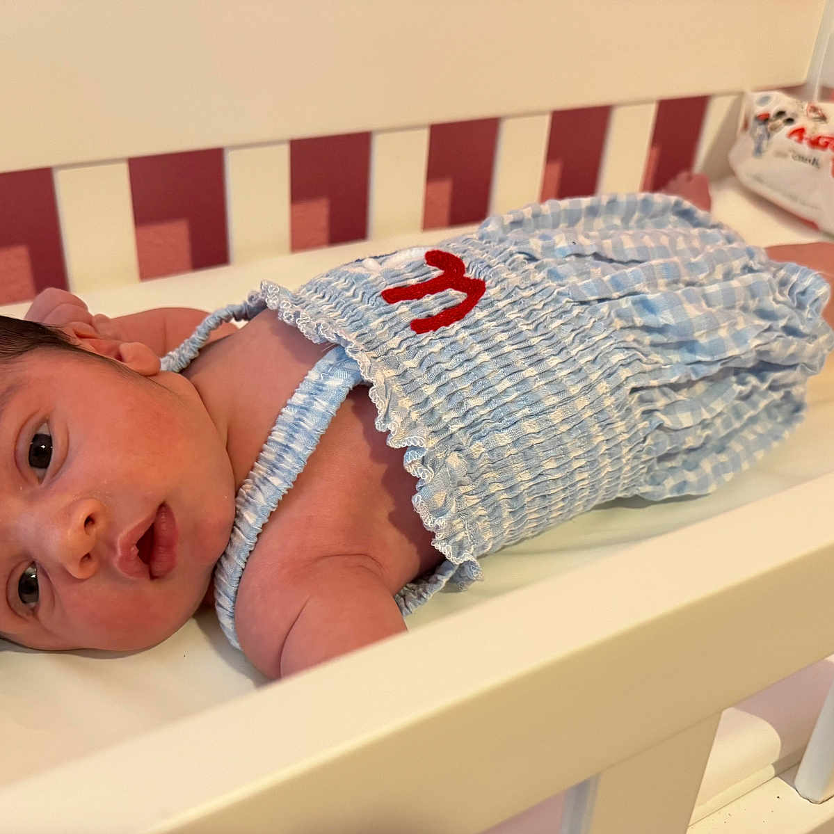 Kylie is registered to the contest to win money with this photo: baby, baby_wipes, blue_clothing, changing_table, child, cute, expression, eyes, face, hair, indoor, infant, lying_down, newborn, portrait, red_embroidery, skin, small, striped_outfit, white_furniture