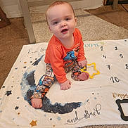 Joshua is registered to the contest to win money with this photo: baby, child, infant, sitting, blanket, toy, dinosaur, orange_clothing, patterned_pants, carpet, indoor, cute, star, numbers, playful, curious, smiling, person, floor, young