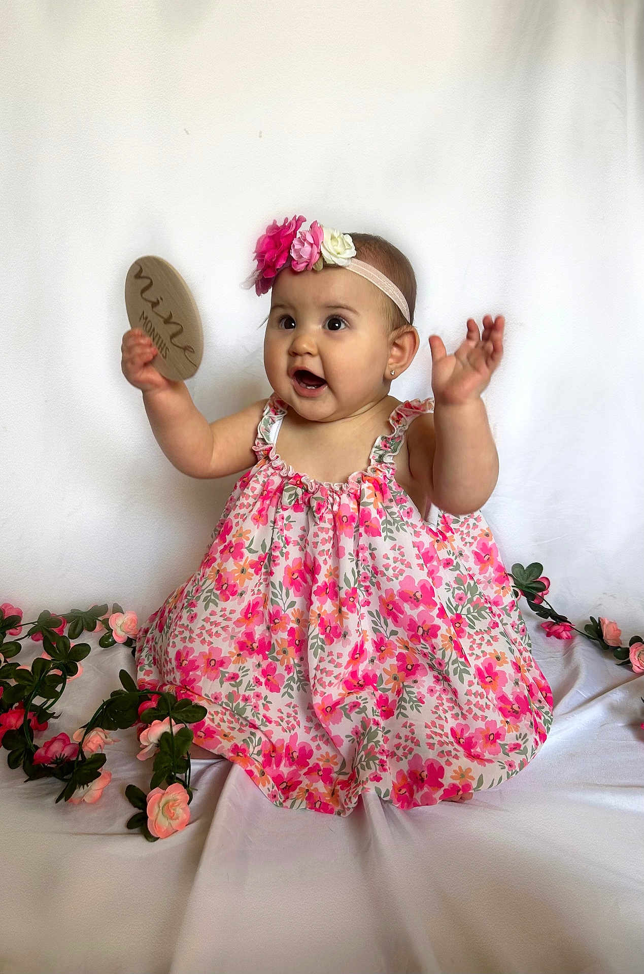 Everly is registered to the contest to win money with this photo: baby, child, girl, floral_dress, flower_headband, pink_flowers, milestone, nine_months, plaque, white_background, smiling, happy, cute, portrait, sitting, decorative_flowers, celebration, hands_up, infant, studio_photo