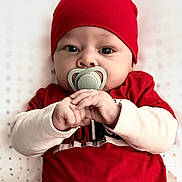Jakari is registered to the contest to win money with this photo: baby, infant, pacifier, red_hat, red_shirt, gray_pants, lying_down, face, hands, cute, child, portrait, baby_clothing, indoor, blanket, phone_screen, lock_screen, time_display, date_display, human