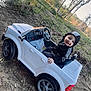 Dawson is registered to the contest to win money with this photo: child, toy_car, outdoor, smiling, hoodie, grass, dirt, trees, pond, nature, play, happy, vehicle, wheel, seat, daylight, person, casual_clothing, young, park