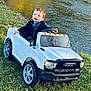 toddler, child, toy_car, outdoor, river, grass, sunlight, happy, blue_eyes, black_jacket, white_toy_truck, nature, daylight, play, vehicle, young_child, smiling, park, water, greenery