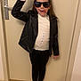 child, leather_jacket, sunglasses, white_shirt, black_pants, pink_socks, indoor, door, floor, playful, tongue_out, hand_sign, standing, expression, clothing_tag, casual, person, hair, rebellious, fun