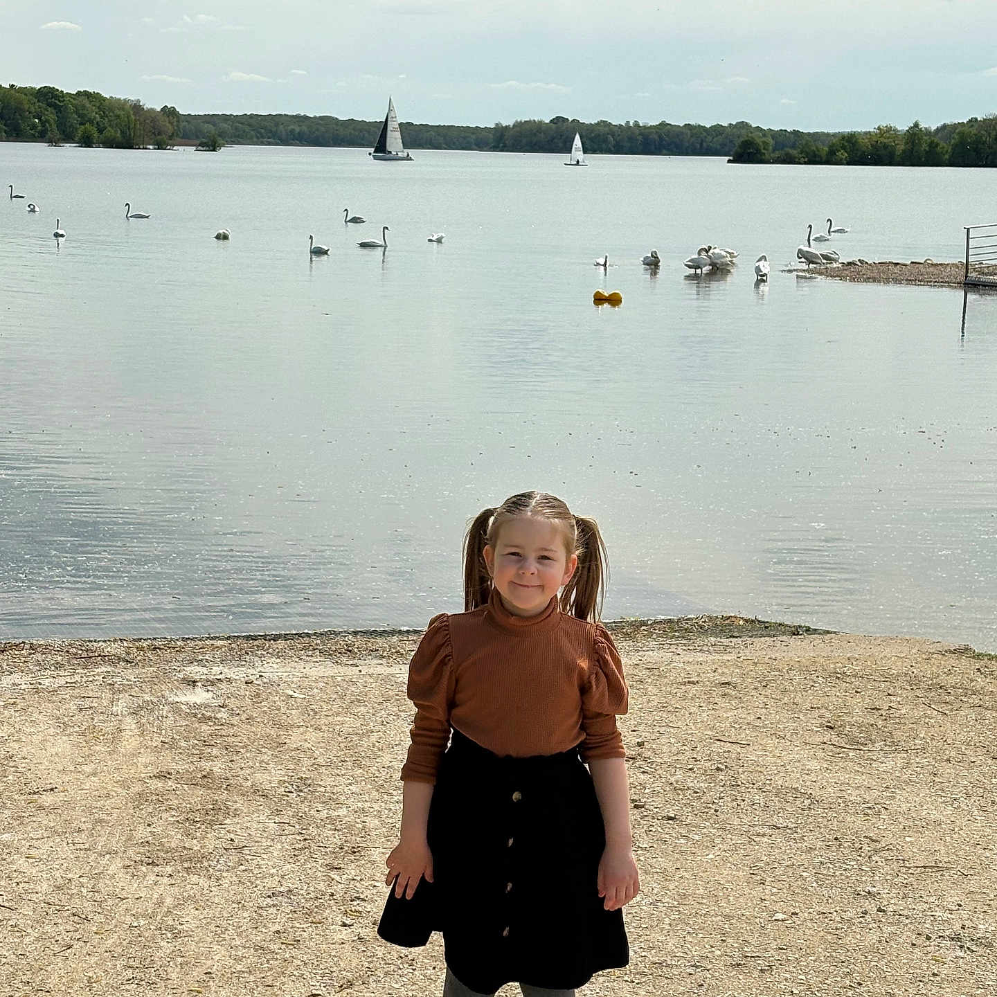 Mia participe au concours pour gagner de l'argent avec cette photo : black_skirt, brown_top, calm, child, cloudy_sky, daytime, girl, gray_tights, lake, nature, outdoor, pigtails, sailboats, sand, shoes, shore, smile, swans, trees, water