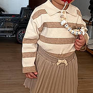 Sofia participe au concours pour gagner de l'argent avec cette photo : accessory, beaded_chain, casual, child, clothing, curious_expression, furniture, home, indoor, pacifier, person, pleated_skirt, sneakers, standing, striped_sweater, tights, toddler, toy_car, wooden_floor, young_child