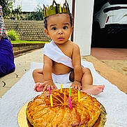 Gabriel participe au concours pour gagner de l'argent avec cette photo : baby, toddler, crown, cake, candles, birthday, outdoor, blanket, sash, diaper, surprised_expression, pastry, golden_plate, garage, car, driveway, garden_wall, sitting, barefoot, celebration