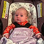 baby, infant, car_seat, blanket, purple_blanket, orange_clothing, floral_pattern, face, head, eyes, hands, safety_harness, indoor, child, newborn, portrait, cute, seated, person, expression