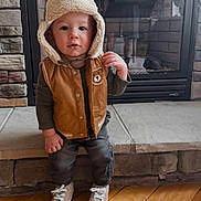 Elijah is registered to the contest to win money with this photo: toddler, child, hat, vest, bear_patch, fireplace, stone_wall, wood_floor, shoes, clothing, indoor, cute, person, portrait, standing, flooring, apparel, warm_clothing, baby, young_child