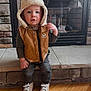 toddler, child, hat, vest, bear_patch, fireplace, stone_wall, wood_floor, shoes, clothing, indoor, cute, person, portrait, standing, flooring, apparel, warm_clothing, baby, young_child