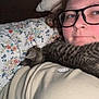 Lenny joined the competition — help win amazing prizes! person, glasses, kitten, cat, lying_down, pillow, floral_pattern, bed, cozy, indoor, pet, animal, tabby, cute, resting, closeup, casual, comfort, sleepy, home
