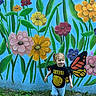 child, toddler, girl, nirvana_tshirt, pigtails, butterfly_wings, mural, flowers, grass, outdoor, playful, colorful, happy, nature, wall_art, casual_clothing, smiling, young_child, blue_background, greenery