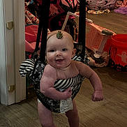 Augustus is registered to the contest to win money with this photo: baby, child, indoor, floor, wood_floor, jumper, smile, face, toy, motorcycle_toy, pink, black_and_white, leaf, halloween_bucket, clothing, person, furniture, playroom, happy, standing