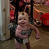 baby, child, indoor, floor, wood_floor, jumper, smile, face, toy, motorcycle_toy, pink, black_and_white, leaf, halloween_bucket, clothing, person, furniture, playroom, happy, standing