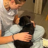 person, puppy, dog, black_dog, pet, sitting, lap, cuddling, stroking, collar, indoors, home, floor, tiles, jeans, white_shirt, puma_logo, tattoo, bracelet, portrait
