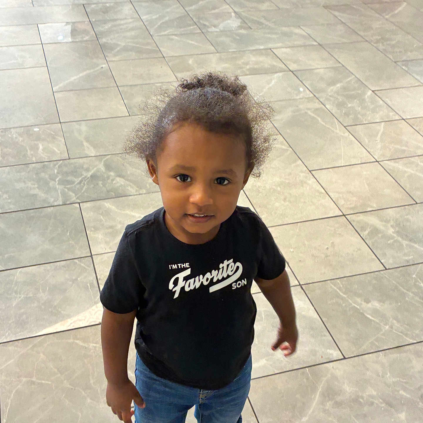 Karter is registered to the contest to win money with this photo: toddler, child, boy, curly_hair, black_tshirt, jeans, blue_crocs, indoor, floor_tiles, standing, looking_up, casual_clothing, cute, young_child, portrait, person, smiling, hair_bun, fashion, playful
