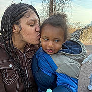 Karter is registered to the contest to win money with this photo: woman, child, kiss, braided_hair, jacket, lollipop, outdoor, pathway, smile, affection, holding, person, nature, daylight, scenic, stone_path, hooded_jacket, emotion, family, casual_clothing