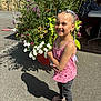 child, girl, barefoot, braids, colorful_hair, pink_top, jeans, flower_pot, flowers, outdoor, sunlight, shadow, smile, pavement, person, elderly, table, chair, greenery, nature