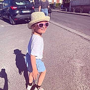 Ilouna a rejoint le concours — aidez-le/la à gagner de superbes lots ! asphalt, blue_shorts, building, car, casual_clothing, child, daylight, footwear, hat, outdoor, person, shadow, sidewalk, smile, street, sunglasses, sunny, urban, white_shirt, woman