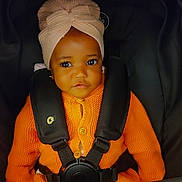 Oluwatobiloba is registered to the contest to win money with this photo: baby, child, infant, car_seat, orange_sweater, head_wrap, turban, seatbelt, cute, portrait, indoors, young_child, face, eyes, clothing, sitting, secure, adorable, warm_clothing, child_seat