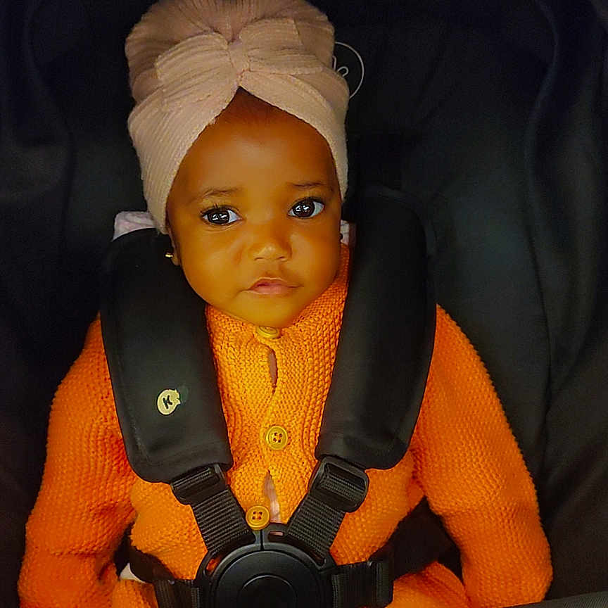 Oluwatobiloba is registered to the contest to win money with this photo: adorable, baby, car_seat, child, child_seat, clothing, cute, eyes, face, head_wrap, indoors, infant, orange_sweater, portrait, seatbelt, secure, sitting, turban, warm_clothing, young_child