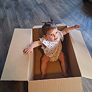 Maïwenne a rejoint le concours — aidez-le/la à gagner de superbes lots ! arms_outstretched, baby, cardboard_box, child, clothing, cute, expression, floor, furniture, home, indoor, infant, light, person, pigtails, playful, room, sitting, wooden_floor, young_child