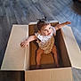 arms_outstretched, baby, cardboard_box, child, clothing, cute, expression, floor, furniture, home, indoor, infant, light, person, pigtails, playful, room, sitting, wooden_floor, young_child