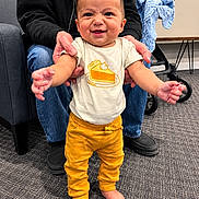 Elijah is registered to the contest to win money with this photo: baby, child, infant, smiling, standing, portrait, indoor, adult, caregiver, support, clothing, yellow_pants, white_shirt, cake_design, bare_feet, blue_blanket, chair, stroller, carpet, happy