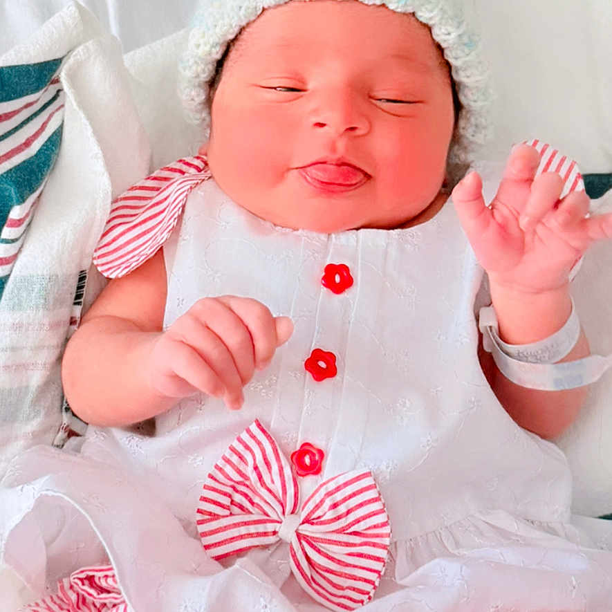 Himaya is registered to the contest to win money with this photo: baby, blanket, bow, button, child, cute, dress, face, hat, hospital_bracelet, indoors, infant, knitted_hat, lying_down, newborn, red_stripes, sleepy, soft_lighting, tongue_out, white_dress