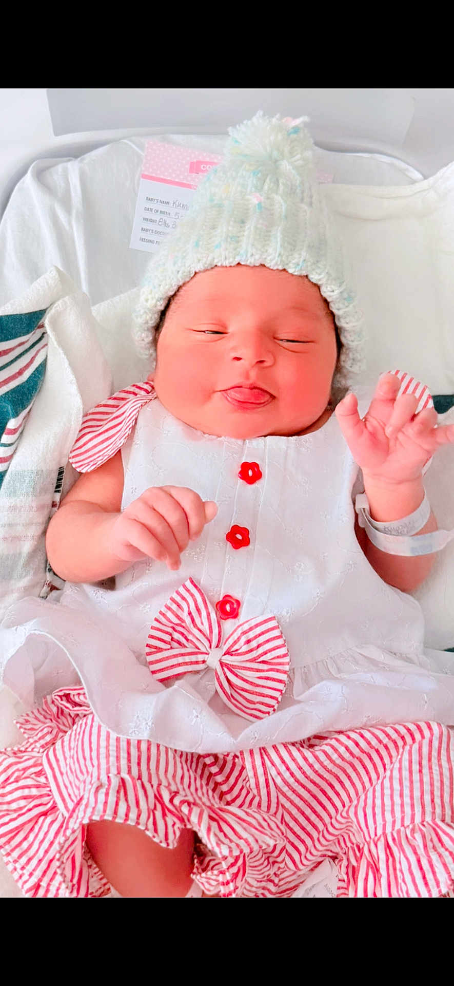 Himaya is registered to the contest to win money with this photo: baby, infant, hat, knitted_hat, dress, white_dress, red_stripes, bow, button, blanket, hospital_bracelet, newborn, cute, face, tongue_out, sleepy, lying_down, soft_lighting, indoors, child