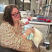 Roselynn joined the competition — help win amazing prizes! woman, baby, newborn, hospital, hospital_room, hospital_bed, medical_equipment, baby_bassinet, floral_gown, glasses, window, snowflake_decorations, chair, pillow, blanket, person, mother, infant, smile, indoor