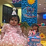 baby, birthday_cake, butterfly_pattern, cake_decorations, cartoon_characters, celebration, child, curly_hair, face, festive, first_birthday, indoor, number_one, party, party_hat, person, pink_dress, smiling, tablecloth, yellow_tablecloth