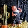 baby, child, infant, plush_cactus, stuffed_plant, denim_overalls, white_shirt, straw, pot, black_background, posing, flexing, cute, smiling, indoors, soft_toys, fabric, portrait, small_arms, playful