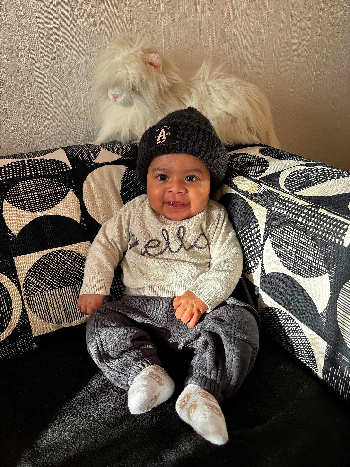 Kenny a rejoint le concours — aidez-le/la à gagner de superbes lots ! baby, child, smiling, hat, knit_hat, plush_toy, stuffed_animal, couch, patterned_cushion, socks, sitting, indoor, happy, cozy, cute, person, portrait, home_interior, soft_toy, warm_clothing