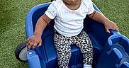 Olivia is registered to the contest to win money with this photo: baby, child, infant, portrait, sitting, ride_on_cart, blue_cart, outdoor, artificial_grass, headband, flower, white_clothing, leopard_print, pants, shoes, serious_expression, looking_at_camera, furniture, play_equipment, photography