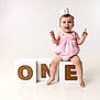 baby, child, one_year_old, birthday, crown, pink_romper, white_blocks, wooden_letters, letter_o, letter_n, letter_e, studio_photo, smiling, happy, celebration, sitting, barefoot, cute, portrait, isolated_background