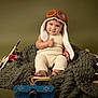 baby, child, pilot_hat, knitwear, toy_airplanes, blanket, smile, thumbs_up, portrait, indoor, cute, infant, vintage_style, playful, cozy, seat, headwear, footwear, red_bracelet, studio